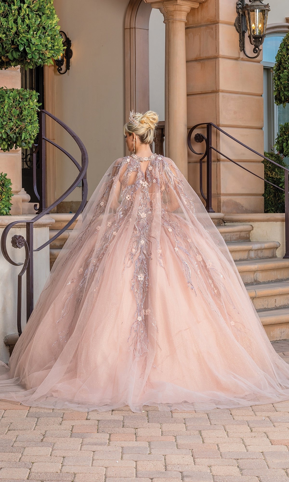 A woman in the Sleeveless Quinceanera Dress 1677, a voluminous blush pink ball gown with a long sheer cape and floral embroidery, stands at the base of stone steps, facing away from the camera.